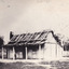 timber homestead with wooden slatted roof and open verandah. A small boy stands on the verandah. A forest can be seen in the background.