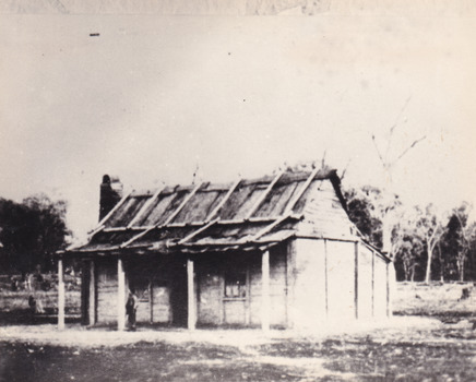 timber homestead with wooden slatted roof and open verandah. A small boy stands on the verandah. A forest can be seen in the background.