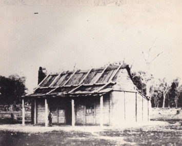 timber homestead with wooden slatted roof and open verandah. A small boy stands on the verandah. A forest can be seen in the background.