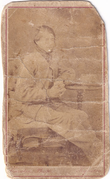 A carte de visits showing a young man (Dan Kelly) with a neck scarf sits on a chair with one elbow leaning on a pedestal side table
