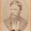 A portrait of light skin tone man, with a long bushy beard, and formally dressed. He is looking off to his right side. 