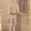 Full shot of man (Aaron Sherritt). He wears a hat, waistcoat and cummerbund and stands with his left arm resting on a podium. A book is placed on the podium.