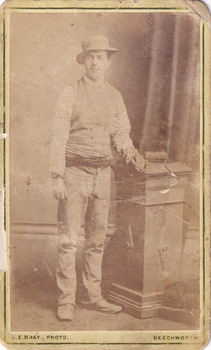 Full shot of man (Aaron Sherritt). He wears a hat, waistcoat and cummerbund and stands with his left arm resting on a podium. A book is placed on the podium.