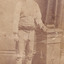 Full shot of man (Aaron Sherritt). He wears a hat, waistcoat and cummerbund and stands with his left arm resting on a podium. A book is placed on the podium.