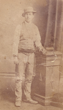 Full shot of man (Aaron Sherritt). He wears a hat, waistcoat and cummerbund and stands with his left arm resting on a podium. A book is placed on the podium.