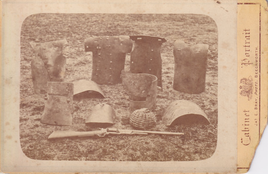 Sepia photograph of Kelly armour including helmets and breastplates arranged on the ground.