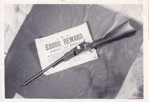 A black and white photograph of a rifle sitting across a poster offering an £8000 reward for robbery and murder. The two objects are resting on a dark material which sits on a shaggy surface. The image has a thin white border.