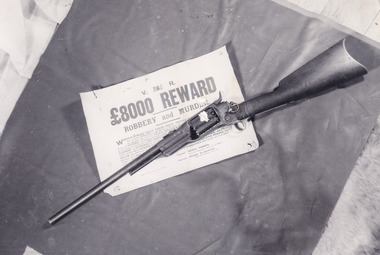 A black and white photograph of a rifle sitting across a poster offering an £8000 reward for robbery and murder. The two objects are resting on a dark material which sits on a shaggy surface.