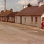 A coloured photograph of a street with four to five cottages, power lines, and two signs (a For Sale sign and a One Way sign). A person in a red dress walks along the footpath, away from the camera.