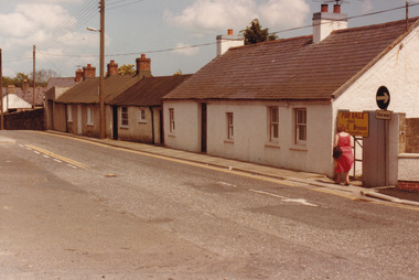 A coloured photograph of a street with four to five cottages, power lines, and two signs (a For Sale sign and a One Way sign). A person in a red dress walks along the footpath, away from the camera.