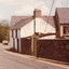 A coloured photograph of a street with two houses pictured and a stone wall. In the background, as the road curves behind the building, four cars are parked on the opposite side of the road.