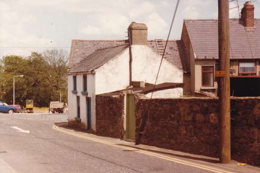 A coloured photograph of a street with two houses pictured and a stone wall. In the background, as the road curves behind the building, four cars are parked on the opposite side of the road.
