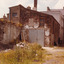 A coloured photograph from the backyard of a large building. Closest to the camera, the building is mostly made of brick with fading paint, while other sections of the building are painted solid colours.