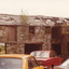 A coloured photograph of a two storey brick building with roof shingles falling off. Three cars are parked in the courtyard created by the brick walls.