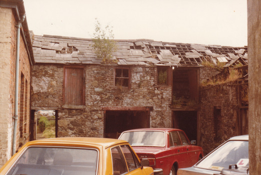 A coloured photograph of a two storey brick building with roof shingles falling off. Three cars are parked in the courtyard created by the brick walls.
