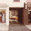 A coloured photograph of a shopfront. On the left is a fashion store, on the right is Burnes Hairdresser. Between the stores is an opening and road, through which a yellow car and old brickwork can be seen.