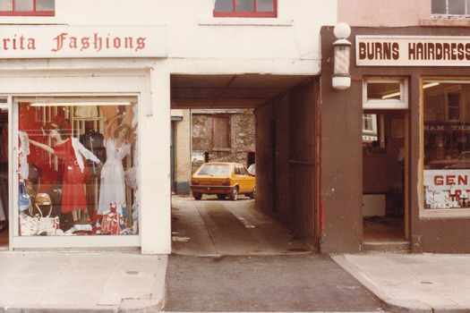 A coloured photograph of a shopfront. On the left is a fashion store, on the right is Burnes Hairdresser. Between the stores is an opening and road, through which a yellow car and old brickwork can be seen.