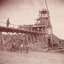Image depicts a massive building with four men near it. One man is on the bridge near the building and the other three are below him. 