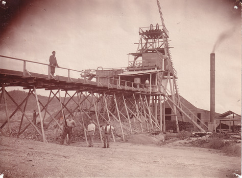 Image depicts a massive building with four men near it. One man is on the bridge near the building and the other three are below him. 