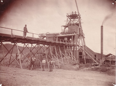 Image depicts a massive building with four men near it. One man is on the bridge near the building and the other three are below him. 