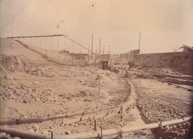Image taken at Allan's flat at one of J.A. Wallace's barge sites. Image depicts water-race and pipes along the ground, connected to two small buildings on high ground.