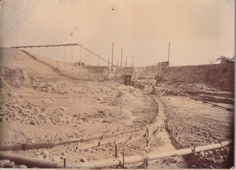 Image taken at Allan's flat at one of J.A. Wallace's barge sites. Image depicts water-race and pipes along the ground, connected to two small buildings on high ground.