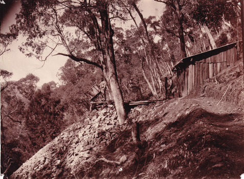 Image taken outside a hut at Beechworth. Image depicts two men standing at the entrance of a wooden hut, with a mine cart near the edge of the cliff. The hut and men are surrounded by trees.