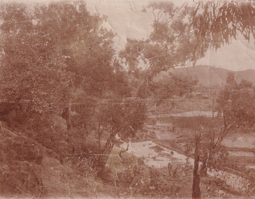 Image taken at Matthew's Gully of a small valley with a building at the base with water surrounding it and a few cottages on the hillside above it. A large portion of the left side of the photos are trees.