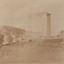 Cropped photograph of a building in the middle ground with a large brick chimney stack to it's right- wood stacks beside it and a hill with trees in the background.
