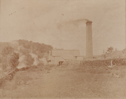 Cropped photograph of a building in the middle ground with a large brick chimney stack to it's right- wood stacks beside it and a hill with trees in the background.