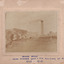 Photograph of a building in the middle ground with a large brick chimney stack to it's right- wood stacks beside it and a hill with trees in the background. Mounted on cardboard with writing (see transcript)