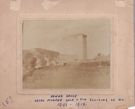Photograph of a building in the middle ground with a large brick chimney stack to it's right- wood stacks beside it and a hill with trees in the background. Mounted on cardboard with writing (see transcript)