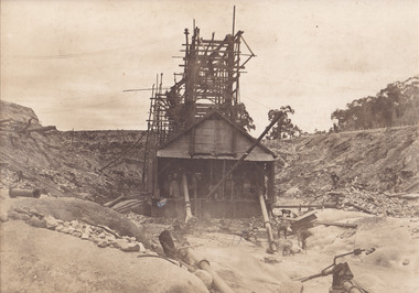 An image taken of Cock's Pioneer  G & T Sluicing No. 4 site looking east. The image depicts several people, including a Mr Hollister (left), Mrs Breustedt (4th from left), the children Min and Chris Breustedt, and Kate Timmons holding a girl's hand, standing underneath the verandah of the timber and tin-roof structure.