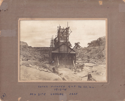 An image with a wide frame taken of Cock's Pioneer  G & T Sluicing No. 4 site looking east. The image depicts several people, including a Mr Hollister (left), Mrs Breustedt (4th from left), the children Min and Chris Breustedt, and Kate Timmons holding a girl's hand, standing underneath the verandah of the timber and tin-roof structure.