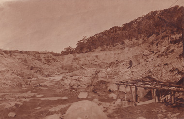 Image taken at El Dorado Cocks Pioneer Gold and Tin Sluicing Company looking east up open cut from barge site. Image depicts unevenly cut rock with a small timber structure on the right of the image