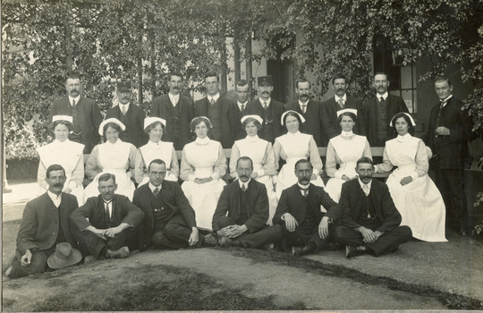Photograph of mental hospital staff, nurses and male workers. 16 men in 2 rows, with a row of female nurses in between. 2 men in the photo have 'Kepi' style hats.