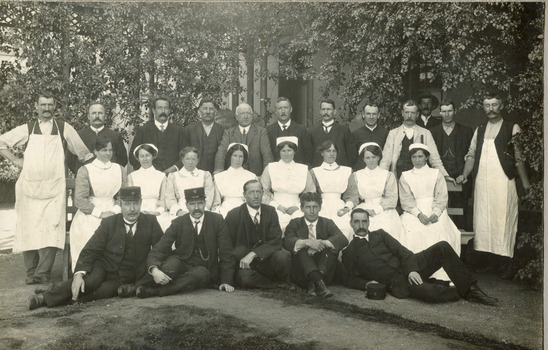 Photograph of mental hospital staff, nurses and male workers. 17 men in 2 rows, with a row of female nurses in between. 2 men in the photo have 'Kepi' style hats.