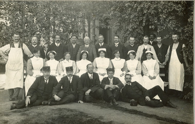 Photograph of mental hospital staff, nurses and male workers. 17 men in 2 rows, with a row of female nurses in between. 2 men in the photo have 'Kepi' style hats.