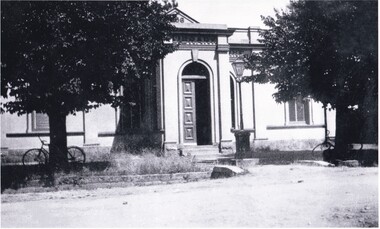 A cropped black and white photograph of the Burke Museum. There are two trees in the foreground which both have bicycles leaning on them.