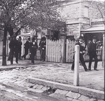 Black and white image of a cobblestone street with a group of men walking in black suits and bowler hats