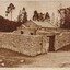 Sepia-toned photograph featuring a small rectangular building with a pitched slate roof and pouch entrance. The building is surrounded by a boundary wall with large pillars and a wooden gate. Both the building and the wall are built of local granite. Trees are in the background.