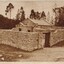 Sepia-toned photograph featuring a small rectangular building with a pitched slate roof and pouch entrance. The building is surrounded by a boundary wall with large pillars and a wooden gate. Both the building and the wall are built of local granite. Trees are in the background.