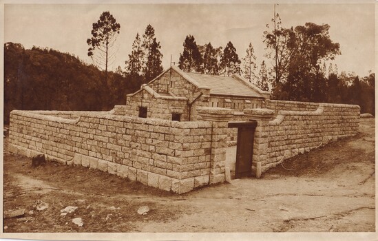 Sepia-toned photograph featuring a small rectangular building with a pitched slate roof and pouch entrance. The building is surrounded by a boundary wall with large pillars and a wooden gate. Both the building and the wall are built of local granite. Trees are in the background.