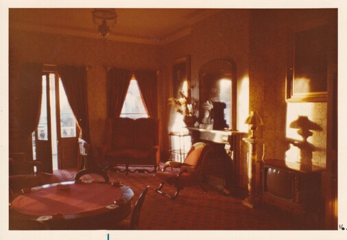 A photograph showing another section of the vintage hotel room featuring another poker table, a  fireplace, tv set and cusioned chairs.