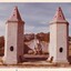 Three woman and a dog posing between two tower like structures with conical tops. In the background is a cemetery with trees lining its edge.