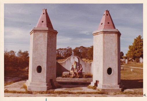 Three woman and a dog posing between two tower like structures with conical tops. In the background is a cemetery with trees lining its edge.