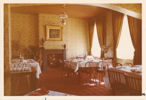 A photograph of a vintage dining room with elaborately laid out tables, a fireplace and a gilded painting hanging above it.