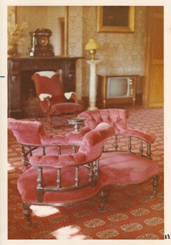 A vintage sitting room with red upholstered furniture, patterned carpet, a fireplace with a clock on the mantel and an old television set.