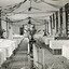 A black and white photograph featuring the internal of a ward in the Mental Hospital. Beds are arranged in two long lines, with a central aisle, and three nurses are in view.