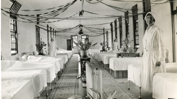 A black and white photograph featuring the internal of a ward in the Mental Hospital. Beds are arranged in two long lines, with a central aisle, and three nurses are in view.